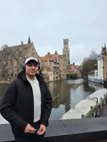 Diego wearing over-ear headphones and a cap, standing by a canal in Bruges with the Belfry tower behind him.