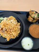 A black tray with a plate of biryani, a bowl of spiced chicken pieces, a bowl of brown curry and a bowl of raita, on a light wooden table.
